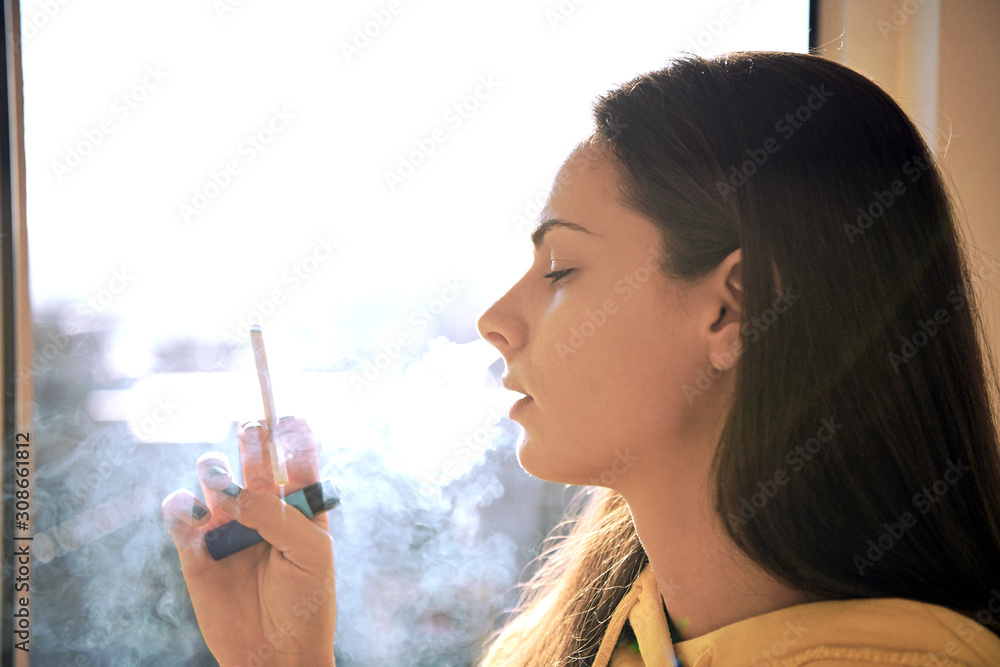 Obraz premium Beautiful attractive girl smoking a cigarette at the window. Pretty woman smoking cigarette.