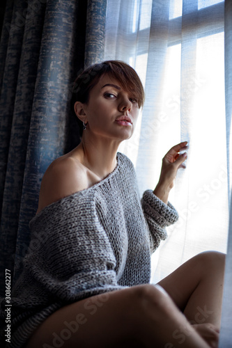 45s years old brunette woman sit on the home floor by the window. Pretty model dressed knitted grey cozy sweater ana look to the window