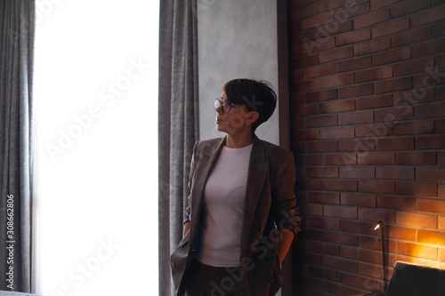  45s years old business woman working at home at the background brick wall , posing against the table with laptop in the evening, take the job at freelance. Entrepreneur wearing a fashion suit