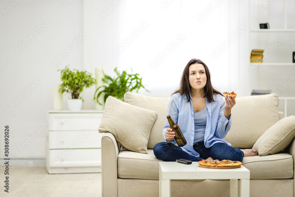 Brunette woman chews, holding a bottle of beer and a slice of pizza hands. Isolated woman on blurred light background. Copy space. Brunette woman chews, holding a bottle of beer and a slice of pizza hands. Isolated woman on blurred light background. Copy space.