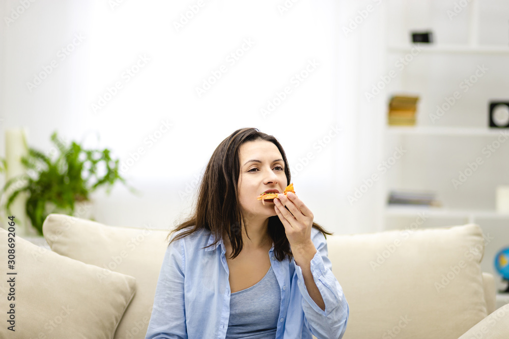 Hungry adorable woman is biting pizza, sitting on the sofa, full of pillows. Close up. Copy space. Hungry adorable woman is biting pizza, sitting on the sofa, full of pillows. Close up. Copy space.
