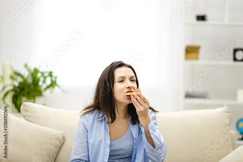 Hungry adorable woman is biting pizza, sitting on the sofa, full of pillows. Close up. Copy space.
