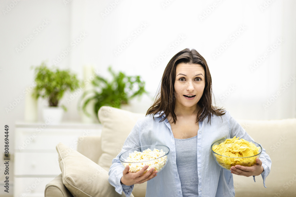 Happy and surprised woman holds deep plates with pop corn and chips.