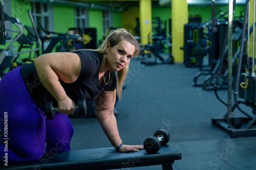 An obese woman is training with dumbbells on a bench in the gym. The fat blonde is losing weight with the help of fitness.
