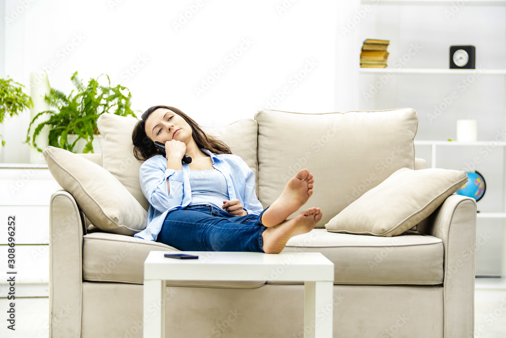 Obraz premium Bored young woman is watching TV, laying on a sofa. Copy space.