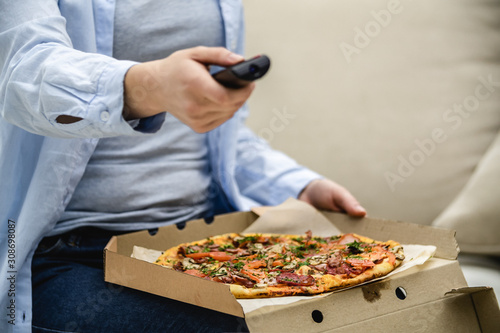 Crop. Unrecognizable woman is sitting on her sofa at home and is watching television while eating pizza. Copy space.
