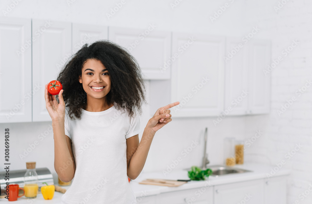 Beautiful smiling African American woman holding tomato, pointing finger on copy space. Healthy lifestyle concept, vegetarian, diet. Attractive emotional girl cooking fresh dinner, standing in kitchen Beautiful smiling African American woman holding tomato, pointing finger on copy space. Healthy lifestyle concept, vegetarian, diet. Attractive emotional girl cooking fresh dinner, standing in kitchen