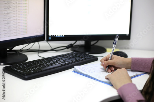 A girl writes in a notebook opposite a computer monitor. Women's hands write. Office worker. Finance and Accounting concept