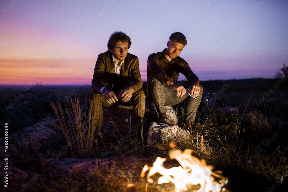 Two Male Friends near Bonfire. Gentlemen has sincere conversation, share memories and drink whiskey. Sunset sky on background