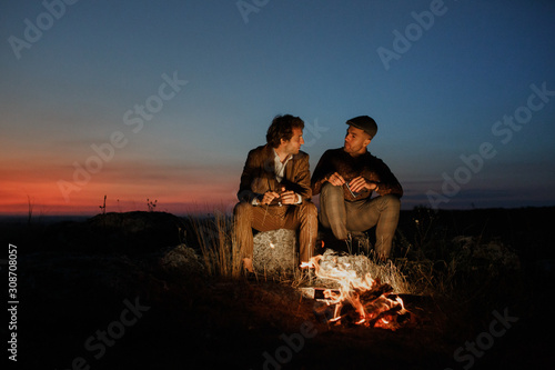 Two Male Friends near Bonfire. Gentlemen has sincere conversation, share memories and drink whiskey. Sunset sky on background