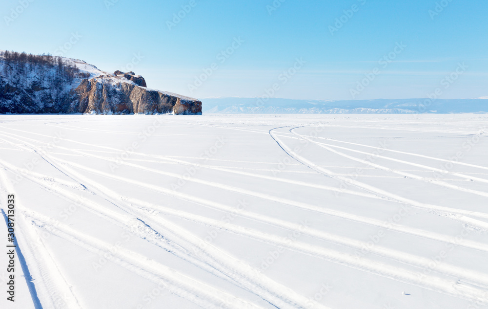 Fototapeta premium Lake Baikal on a sunny winter day. Car tracks on the snowy ice of a lake. Near the cliff, a group of tourists on a picnic. Winter ice travel concept