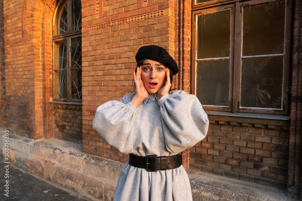 Scared young woman in black beret and grey dress holds her hands near her face and looking at the camera, red brick wall with old wooden window on the background Scared young woman in black beret and grey dress holds her hands near her face and looking at the camera, red brick wall with old wooden window on the background