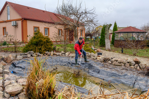 man cleans a garden pond with a landing net from slime, falling leaves and catches fish before winter