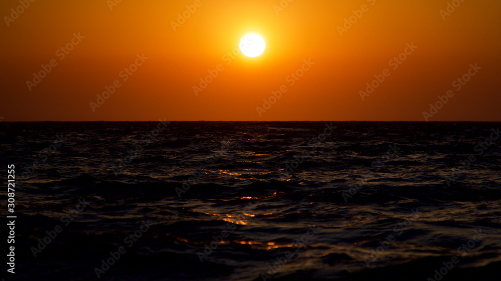 Spectacular sunset with endless horizon and sparkling waves. Lighthouse and rocky hills in the background.