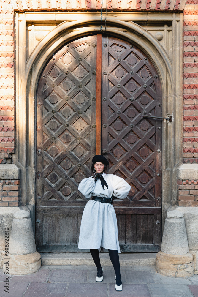 Fashionable young female in black beret and grey old-fashioned dress holds one hand on her waist other hand ner her face and seriously looking at the camera, old vintage wooden door on the background