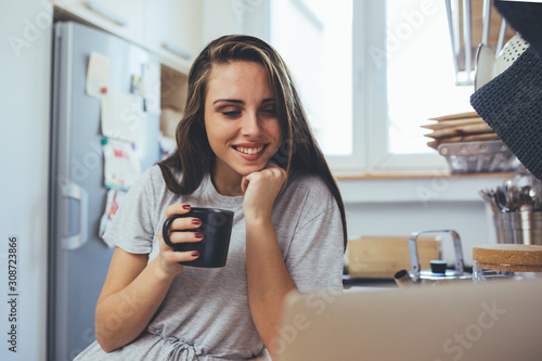 woman using laptop computer and drinking coffee in kitchen on kitchen counter