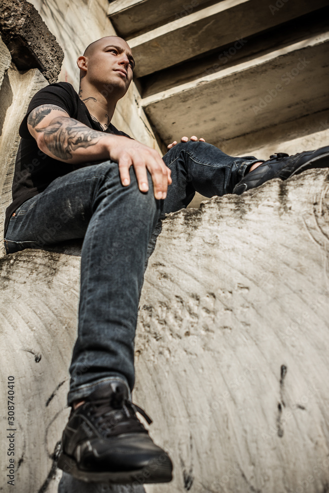 Young attractive stern guy in tattoos sits on a window frame of a destroyed building. Brutal young man with tattoos on his arms and neck sits on the wall of an abandoned house. Bully, bad guy.