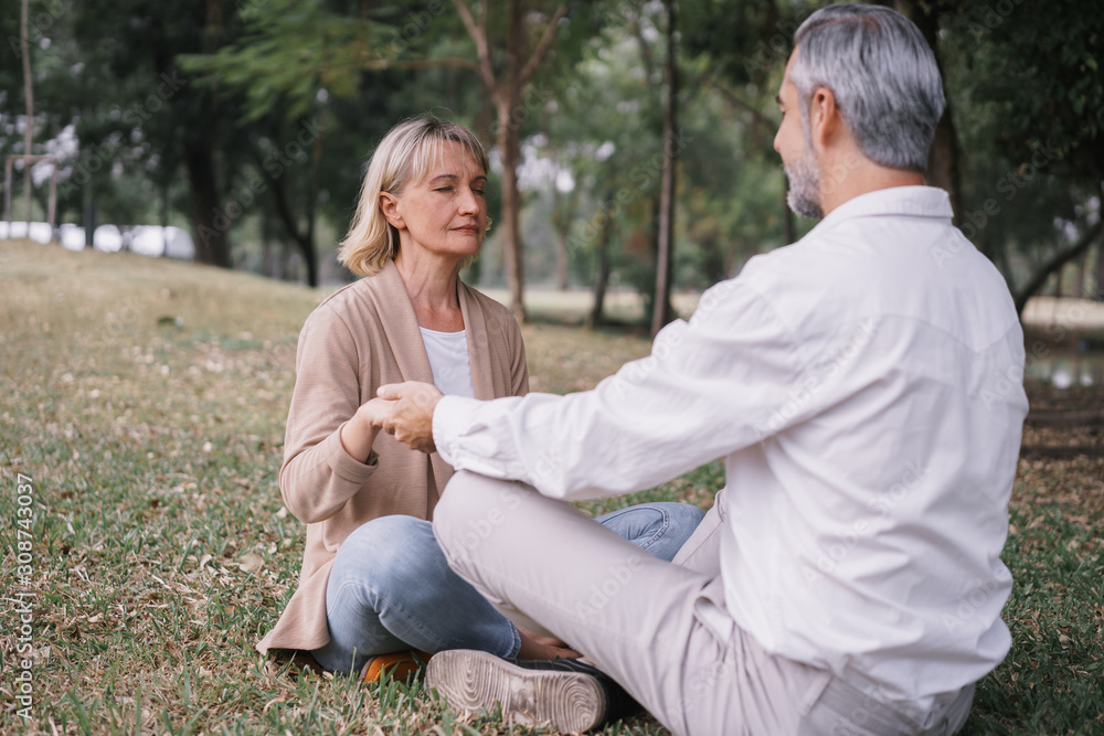Fototapeta premium Elderly couple in practicing yoga while sitting holding to hand together in the public park. Concept of relaxation and meditation.
