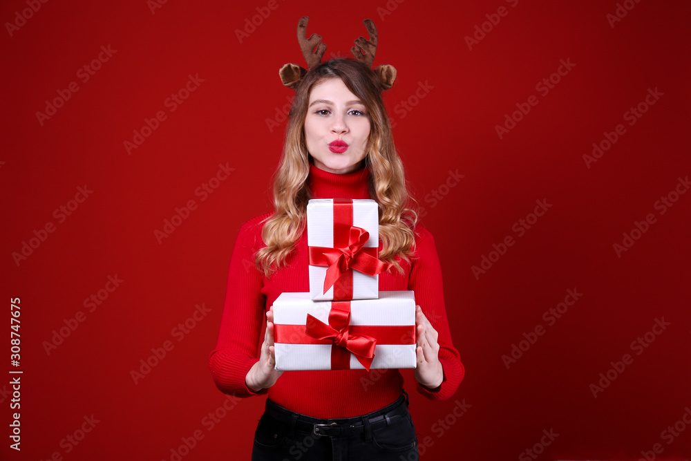Fototapeta premium Studio shot of beautiful smiling young woman wearing reindeer horns headband for Christmas. Attractive blonde female in red turtleneck sweater, new years eve. Copy space, isolated background, close up