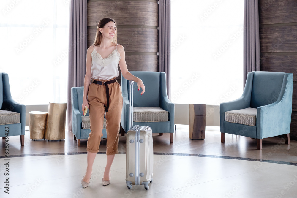 Pretty young smiling woman with smartphone and with suitcase stands in the hotel lobby during check-in. Travel concept and good service. Online booking. Business trip, official journey, business visit
