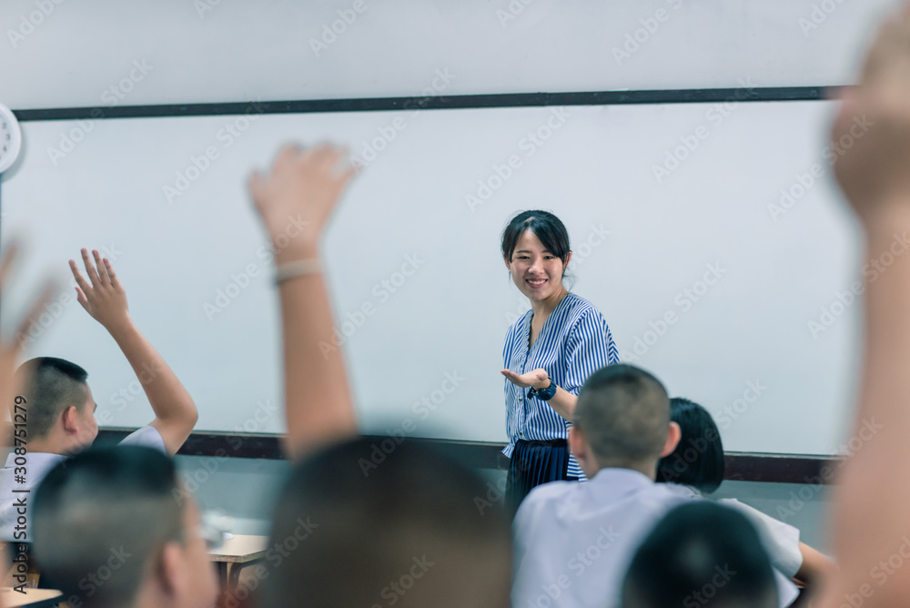 A smiling Asian female high school teacher teaches the white uniform students in the classroom by asking questions and then the students raise their hands for answers. A smiling Asian female high school teacher teaches the white uniform students in the classroom by asking questions and then the students raise their hands for answers.