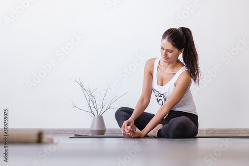 Beautiful well-groomed young brunette woman doing foot massage while sitting on a rug on the floor after yoga classes at home. Concept of healing and self-massage. Copyspace