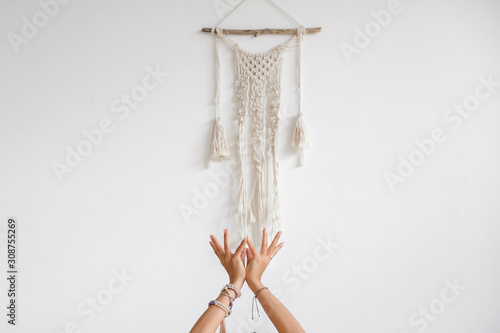 Hands of an unidentified young woman yoga teacher with beautiful bracelets while performing asanas against a white wall and macrame knitted pattern. Yoga and pilates concept