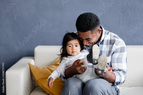 Caring mixed race father holds his pretty little daughter in his arms and puts on her shoe while sitting in a cozy sofa in the living room. Concept of love between father and daughter. Copyspace