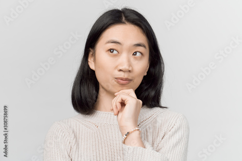 Girl looks up, purses lips, holding fist on chin. Businesslike young woman Asian appearance with black hair and brown eyes dressed in knitted warm sweater stands isolated white background in Studio. © EverGrump