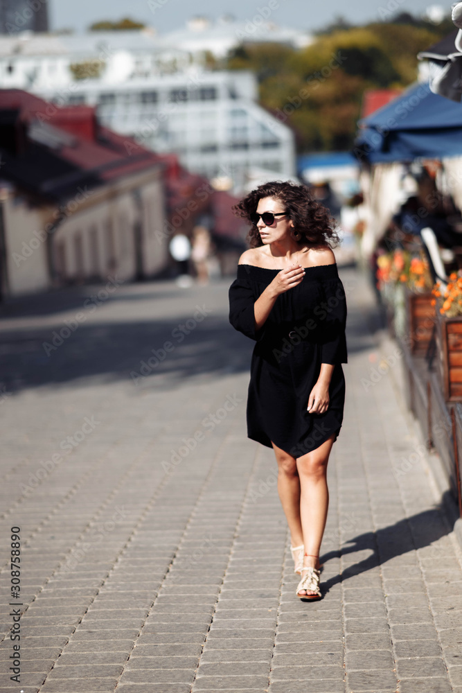 Beautiful curly brunette woman in sunglasses and a black dress walks around the city on a bright sunny day.