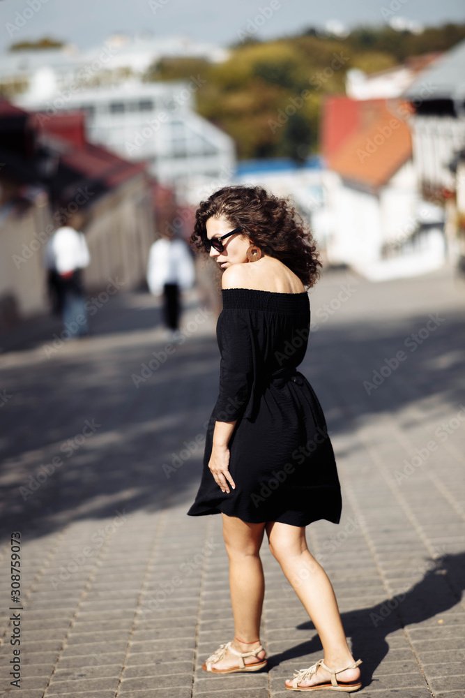 Beautiful curly brunette woman in sunglasses and a black dress walks around the city on a bright sunny day.