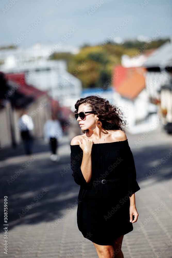 Beautiful curly brunette woman in sunglasses and a black dress walks around the city on a bright sunny day.