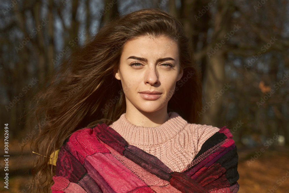 A feminine lady frowns from the sun. Portrait of young woman with brown hair, green eyes wearing pink sweater walking in colorful forest or Park. Girl looking into camera has natural beauty. A feminine lady frowns from the sun. Portrait of young woman with brown hair, green eyes wearing pink sweater walking in colorful forest or Park. Girl looking into camera has natural beauty.