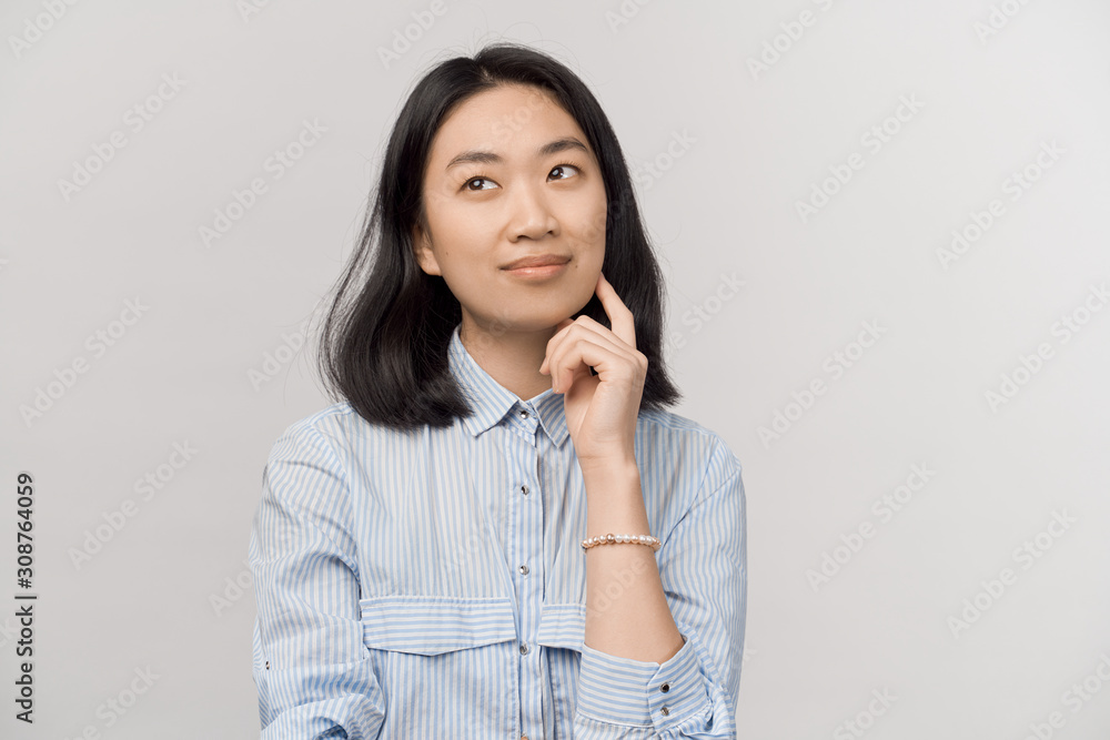 Girl thoughtfully looks up blank space for text. Businesslike beautiful young woman of Asian appearance with black hair dressed in striped office shirt stands isolated white background in Studio. Girl thoughtfully looks up blank space for text. Businesslike beautiful young woman of Asian appearance with black hair dressed in striped office shirt stands isolated white background in Studio.