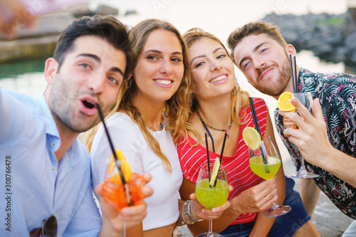 Group of best friends having fun making selfies with smartphone and drinking colorful cocktails at the seaside