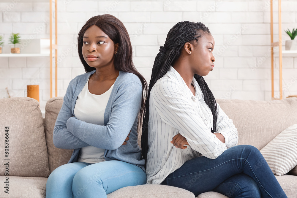 Two offended afro girlfriends sitting back to back on sofa - obrazy, fototapety, plakaty 