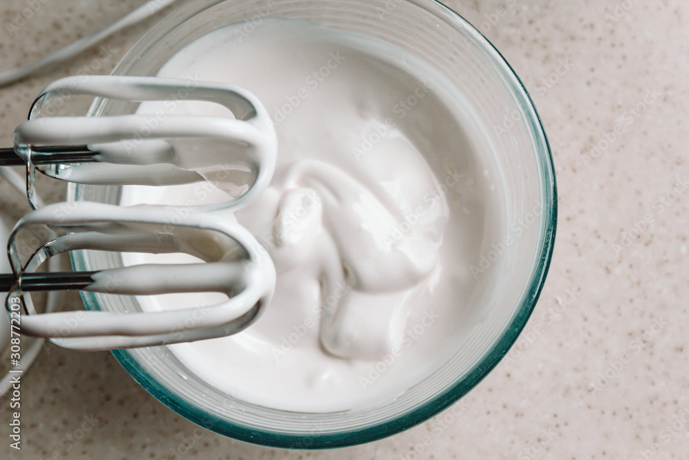 Baking concept. Whipped cream of egg whites for perfect peaks in a glass bowl, with a mixer on a marble white table. Meringue cookie recipe. Baking concept. Whipped cream of egg whites for perfect peaks in a glass bowl, with a mixer on a marble white table. Meringue cookie recipe.
