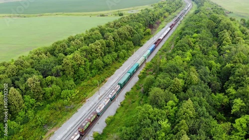 Aerial shot of freight train that travels by rail. Train carries goods and raw materials for heavy industry. Railways are surrounded by trees and endless green fields. Taken by drone at sunset