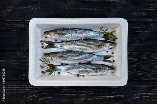 Photography Raw mullet fish with spices and herbs ingredients in white ceramic backing dish on black wooden table ready for cooking
