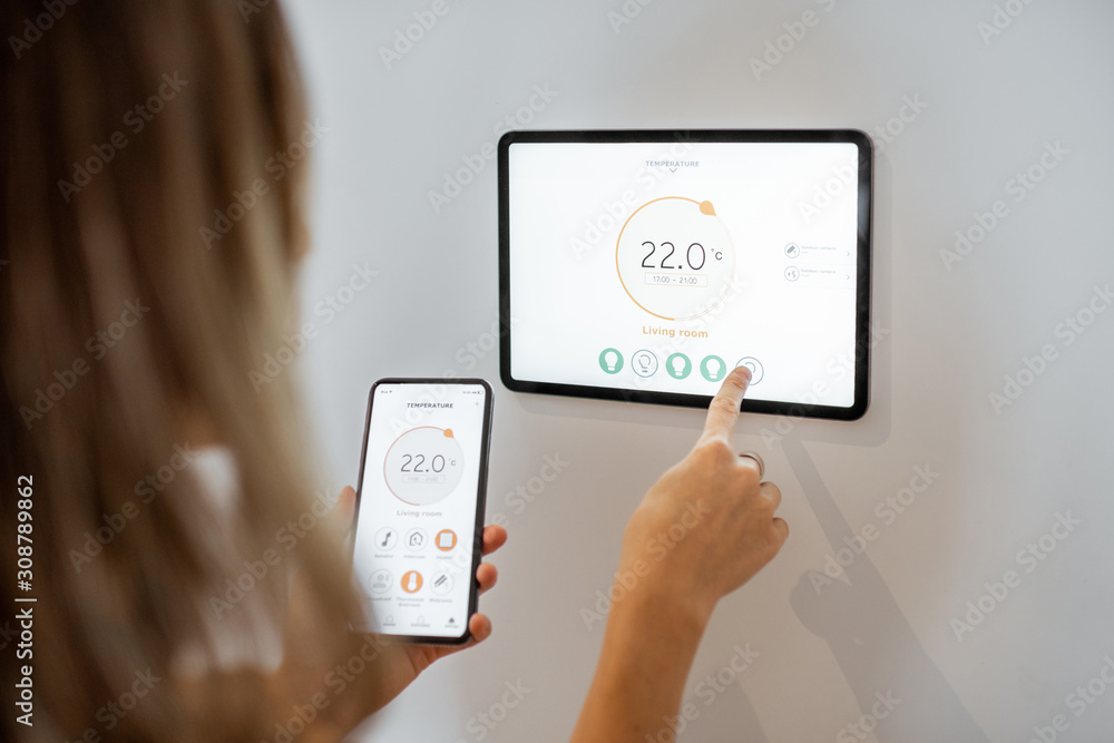 © rh2010 - Young woman controlling temperature in the living room with smart phone and digital touch screen panel. Concept of heating control in a smart home © rh2010 - Young woman controlling temperature in the living room with smart phone and digital touch screen panel. Concept of heating control in a smart home