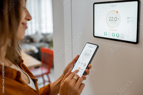 Young woman controlling temperature in the living room with smart phone and digital touch screen panel. Concept of heating control in a smart home photo