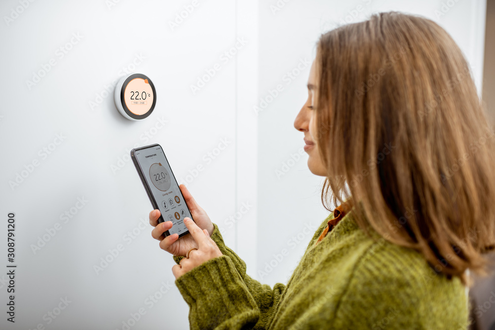 Woman dressed in green sweater regulating heating temperature with a modern wireless thermostat and smart phone at home. Synchronization of thermostat with mobile devices concept - obrazy, fototapety, plakaty 