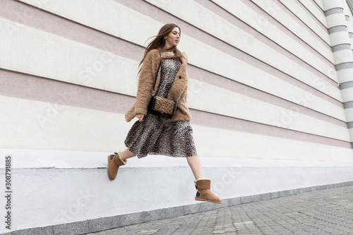 brown fur coat faux fur, long leopard print dress, warm uggs, shoulder bag, beautiful European model girl with long hair posing fun outdoors near the wall photo