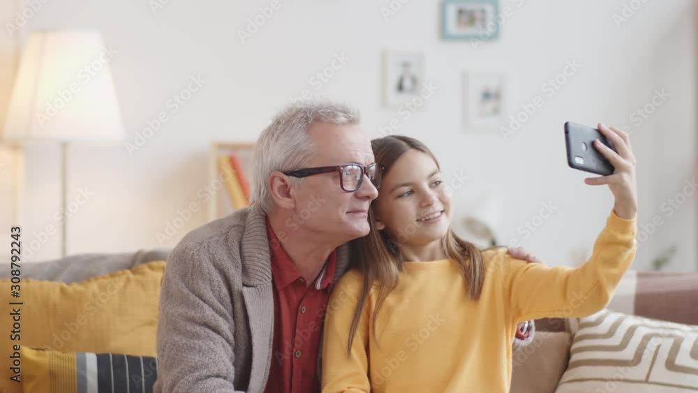 Chest-up shot of smiling 10-year-old Caucasian granddaughter and senior grandfather sitting together on couch at home, girl holding smartphone and taking selfies, then looking at photos on screen Chest-up shot of smiling 10-year-old Caucasian granddaughter and senior grandfather sitting together on couch at home, girl holding smartphone and taking selfies, then looking at photos on screen