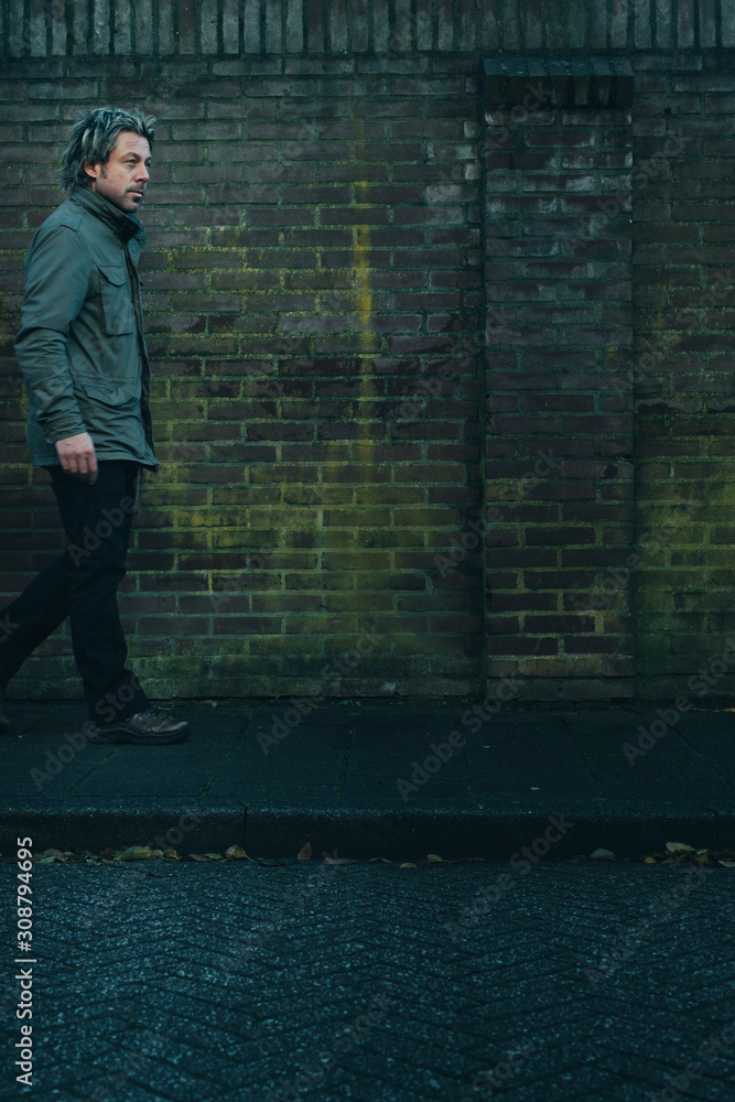 Man in green jacket walking on pavement along brick wall. Man in green jacket walking on pavement along brick wall.