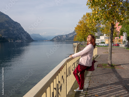 Smiling woman holds grab bar at the lake and looks towards the sun. Autumn day in Como, Italy