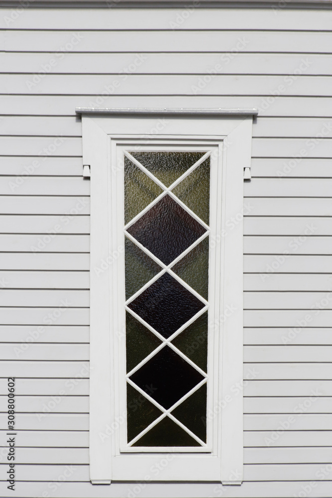 Fototapeta premium Window of traditional wooden rural house. Abstract village architecture fragment featuring narrow stained glass, triangular and rhomboid frames and white wood siding or wall cladding.