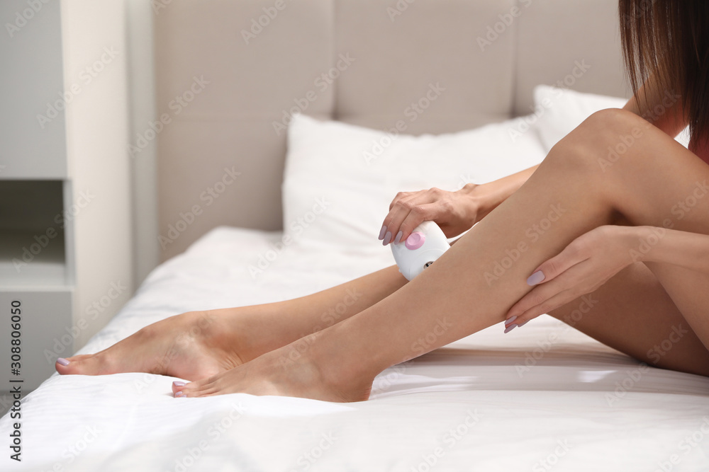 Fototapeta premium Young woman epilating her legs in bedroom, closeup