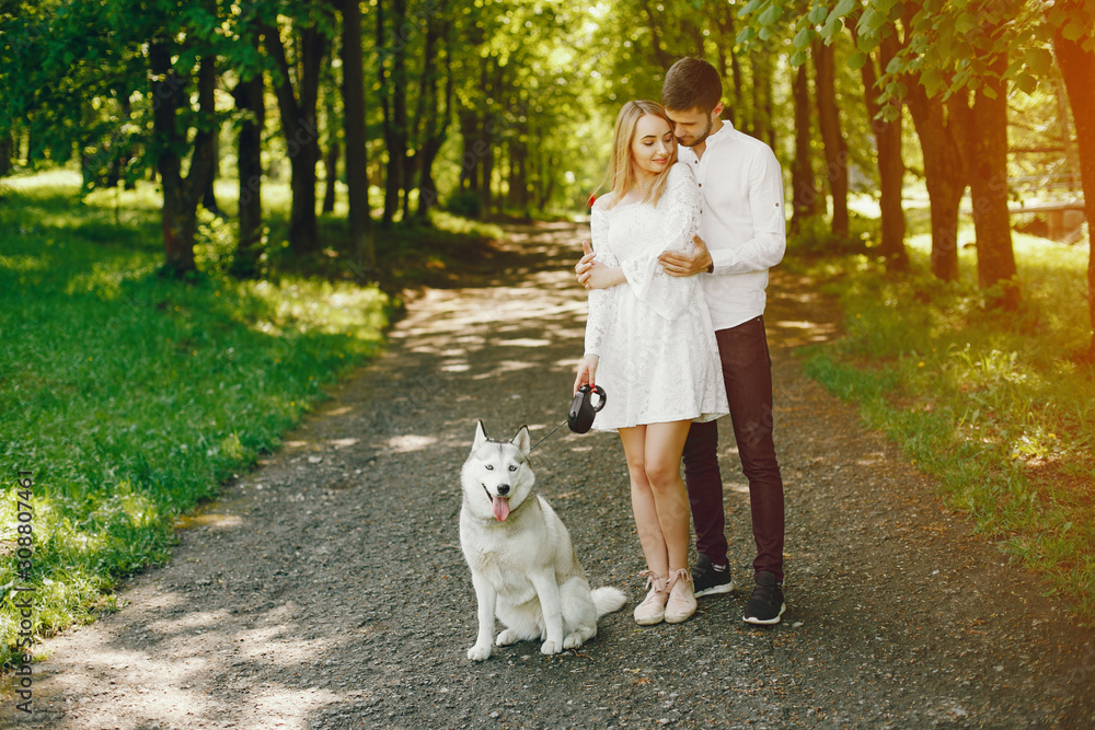 A beautiful and gentle girl with light hair and a white dress is walking in a sunny summer forest with her handsome guy in a white shirt and dark pants and they playing with cute gray dog A beautiful and gentle girl with light hair and a white dress is walking in a sunny summer forest with her handsome guy in a white shirt and dark pants and they playing with cute gray dog