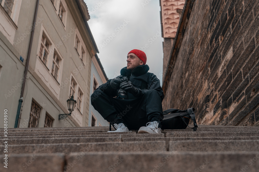 Attractive Man Sitting on Stairs Steps in the Center Of Old European City And Holding Photo Camera. Contemporary Stylish Blogger And Photographer.